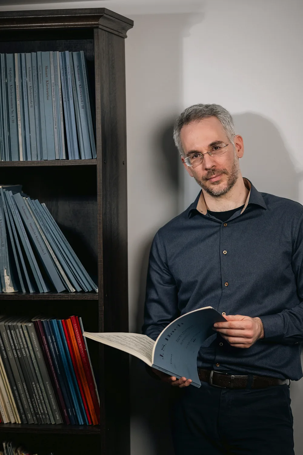 Dimitris Petrovas standing beside his score library, holding a Bach edition